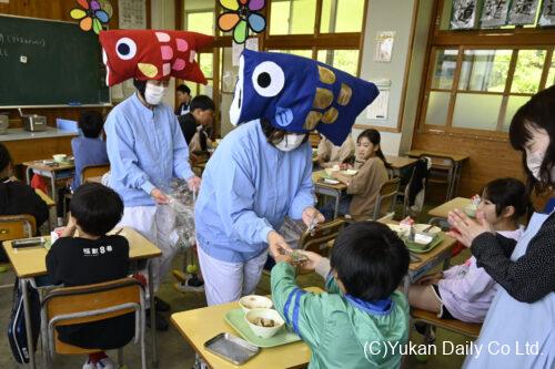 こいのぼりのかぶり物をして児童にかしわ餅を配る給食調理員